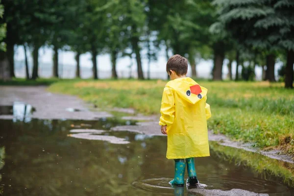 雨の夏の公園で遊んでいる男の子 雨の中で水たまりや泥の中でジャンプ傘 防水コートやブーツと子供 子供は夏の雨の中を歩くどんな天気でも屋外の楽しみ 幸せな子供時代 — ストック写真