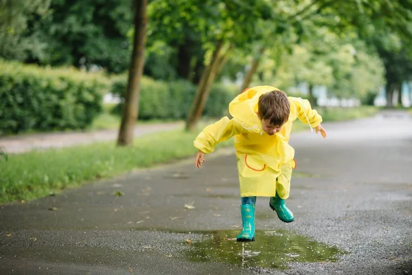 雨の夏の公園で遊んでいる男の子 雨の中で水たまりや泥の中でジャンプ傘 防水コートやブーツと子供 子供は夏の雨の中を歩くどんな天気でも屋外の楽しみ 幸せな子供時代 — ストック写真