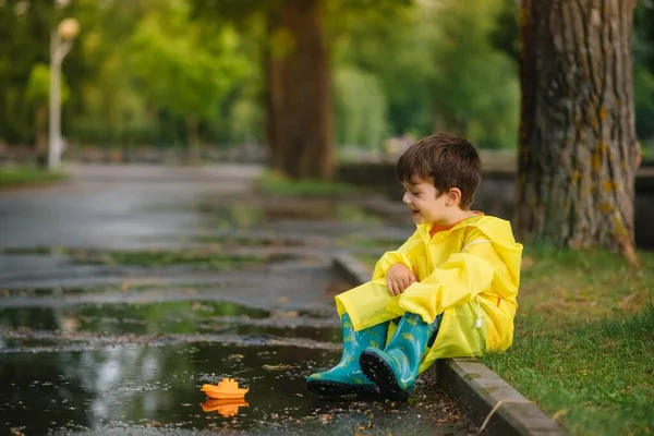 子供は水たまりでおもちゃのボートで遊ぶ 子供は雨で屋外で遊ぶ 子供のための秋の雨の天気屋外活動 泥だらけの水たまりに飛び込む子供 赤ちゃんのための防水ジャケットとブーツ 夏休み 幸せな子供時代 — ストック写真