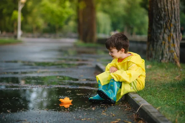 子供は水たまりでおもちゃのボートで遊ぶ 子供は雨で屋外で遊ぶ 子供のための秋の雨の天気屋外活動 泥だらけの水たまりに飛び込む子供 赤ちゃんのための防水ジャケットとブーツ 夏休み 幸せな子供時代 — ストック写真