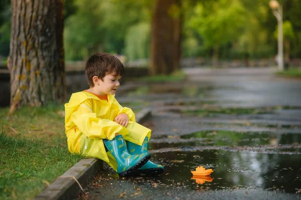子供は水たまりでおもちゃのボートで遊ぶ 子供は雨で屋外で遊ぶ 子供のための秋の雨の天気屋外活動 泥だらけの水たまりに飛び込む子供 赤ちゃんのための防水ジャケットとブーツ 夏休み 幸せな子供時代 — ストック写真