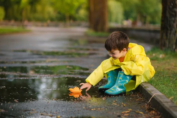 子供は水たまりでおもちゃのボートで遊ぶ 子供は雨で屋外で遊ぶ 子供のための秋の雨の天気屋外活動 泥だらけの水たまりに飛び込む子供 赤ちゃんのための防水ジャケットとブーツ 夏休み 幸せな子供時代 — ストック写真