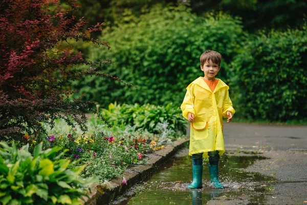 Little boy playing in rainy summer park. Child with umbrella, waterproof coat and boots jumping in puddle and mud in the rain. Kid walking in summer rain Outdoor fun by any weather. happy childhood