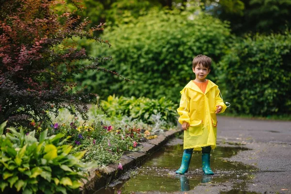 雨の夏の公園で遊んでいる男の子 雨の中で水たまりや泥の中でジャンプ傘 防水コートやブーツと子供 子供は夏の雨の中を歩くどんな天気でも屋外の楽しみ 幸せな子供時代 — ストック写真