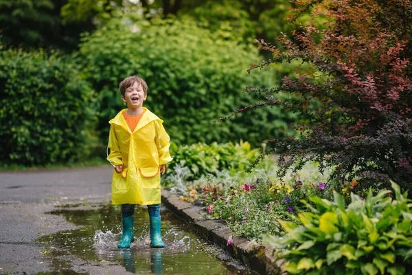 雨の夏の公園で遊んでいる男の子 雨の中で水たまりや泥の中でジャンプ傘 防水コートやブーツと子供 子供は夏の雨の中を歩くどんな天気でも屋外の楽しみ 幸せな子供時代 — ストック写真