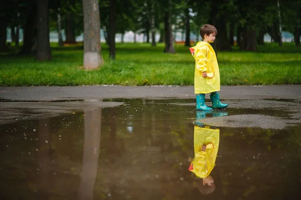 雨の夏の公園で遊んでいる男の子 雨の中で水たまりや泥の中でジャンプ傘 防水コートやブーツと子供 子供は夏の雨の中を歩くどんな天気でも屋外の楽しみ 幸せな子供時代 — ストック写真