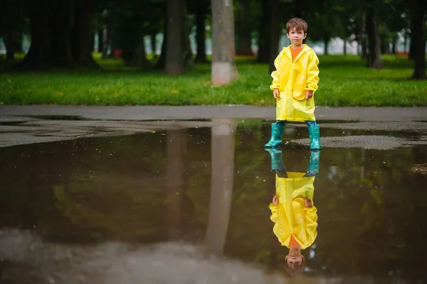 雨の夏の公園で遊んでいる男の子 雨の中で水たまりや泥の中でジャンプ傘 防水コートやブーツと子供 子供は夏の雨の中を歩くどんな天気でも屋外の楽しみ 幸せな子供時代 — ストック写真