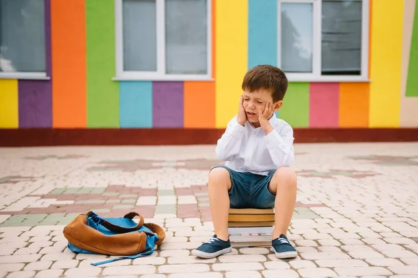 Sad little boy outside of school. Sad schoolboy with books near a modern school. School concept. Back to school.