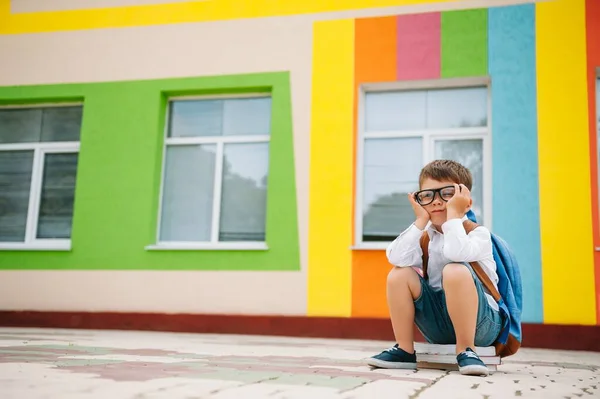 学校の外で悲しい少年 現代の学校の近くに本を持つ悲しい小学生 学校のコンセプト 学校に戻る — ストック写真