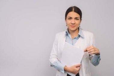 Beyaz üniformalı kadın doktor beyaz arka planda dikilirken elinde mataralar tutuyor..
