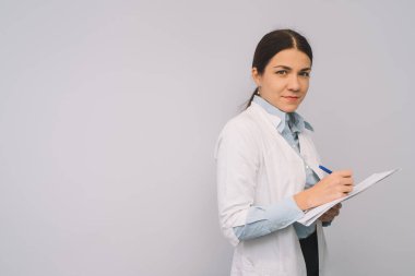 Beyaz üniformalı kadın doktor beyaz arka planda dikilirken elinde mataralar tutuyor..