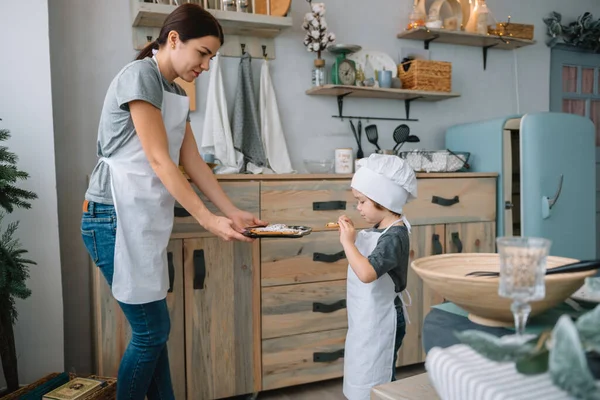 Genç mutlu anne ve bebeği mutfakta kurabiye pişiriyor. Noel ev yapımı zencefilli çörek. Beyaz üniformalı, şapkalı, çikolatalı kurabiyeli tatlı çocuk.