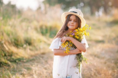 Açık havada, bronzlaşmış sağlıklı tenli, beyaz elbiseli ve yaz şapkalı hoş görünümlü genç bir kız, park yerinde kendinden emin bir ifadeyle poz veriyor, eğlenmeyi seviyor. Güzel genç bayan.