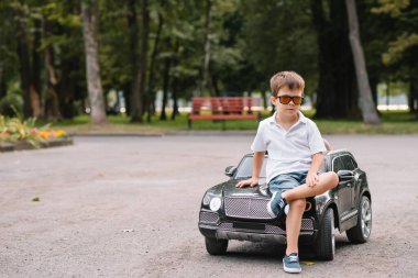 Parkta siyah elektrikli araba süren sevimli çocuk. Komik çocuk elektrikli bir oyuncak arabaya biniyor. Parkta siyah elektrikli araba süren küçük mutlu çocuk. Boşluğu kopyala.