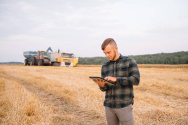 Buğday tarlasında dizüstü bilgisayarı olan genç ve çekici bir çiftçi ve arka planda hasat makinası.