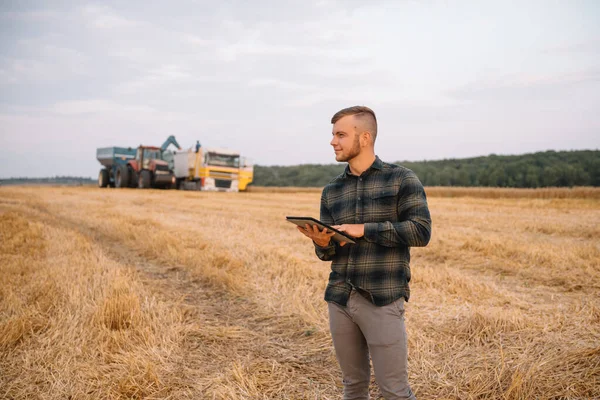 小麦畑にノートパソコンを持つ若い魅力的な農家は 背景に収穫者を組み合わせる — ストック写真