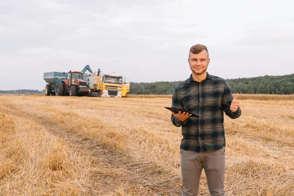 年轻的农艺师站在小麦田里一边 一边检查庄稼的品质 一边联合收割机工作 — 图库照片