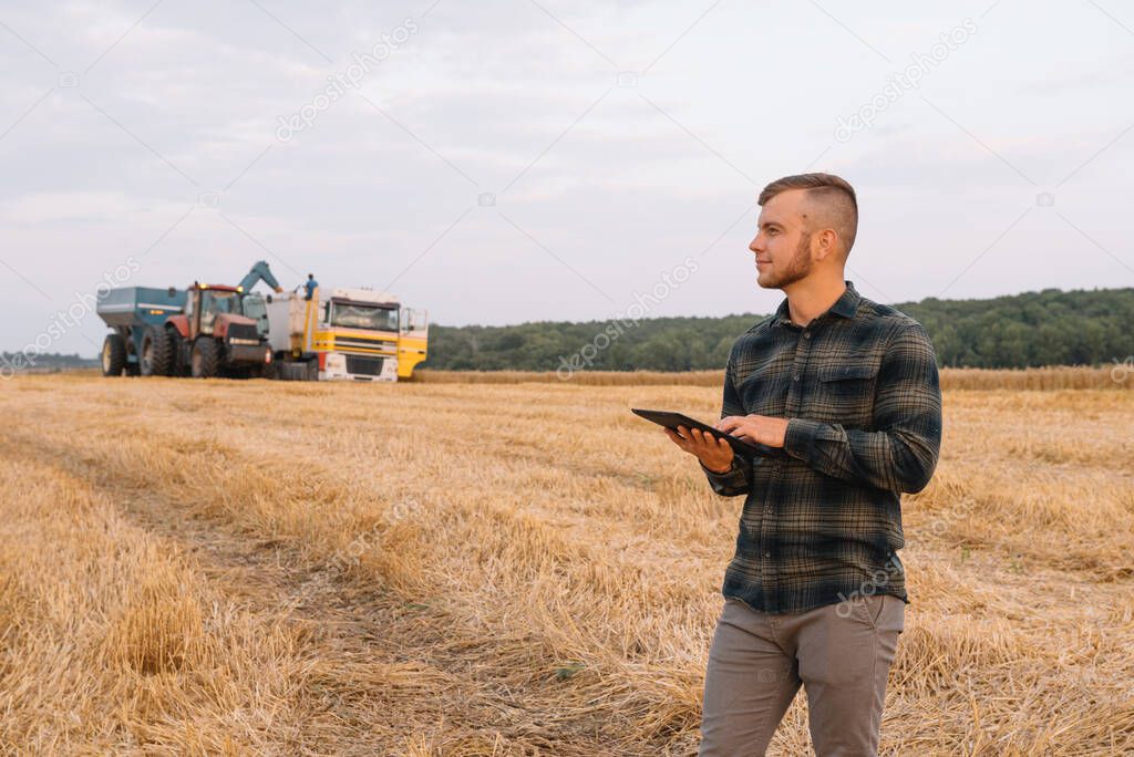 Happy young farmer engineer with notebook standing on wheat field while combine harvester working in background