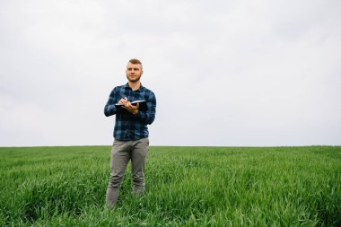 Genç tarımcı yeşil buğday tarlasında defter tutuyor. Tarım sektörü konsepti. Yazın elinde tabletle bir buğday tarlasında duran tarım mühendisi