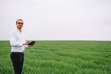 Genç tarım uzmanının elinde yeşil buğday tarlasında tablet dokunmatik tablet bilgisayarı var. Tarım sektörü konsepti. Yazın elinde tabletle bir buğday tarlasında duran tarım mühendisi