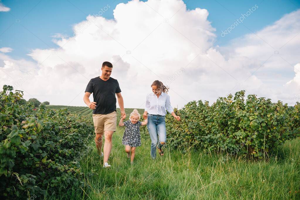 Happy family walking in the park. Mom, dad and daughter walk outdoors, parents holding the baby girl's hands. Childhood, parenthood, family bonds, marriage concept