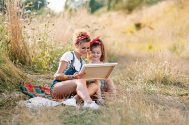 Genç çekici anne, kızına yaz parkında resim yapmayı öğretiyor. Okul çağındaki çocuklar için açık hava aktivitesi