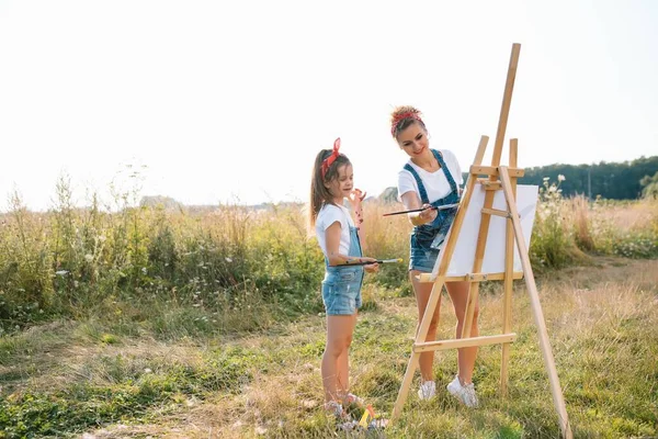 美人の母は娘と絵を描く 女の子と一緒に絵を描くスタイリッシュな女性 白いTシャツと青いジーンズのかわいい子供 母の日 — ストック写真