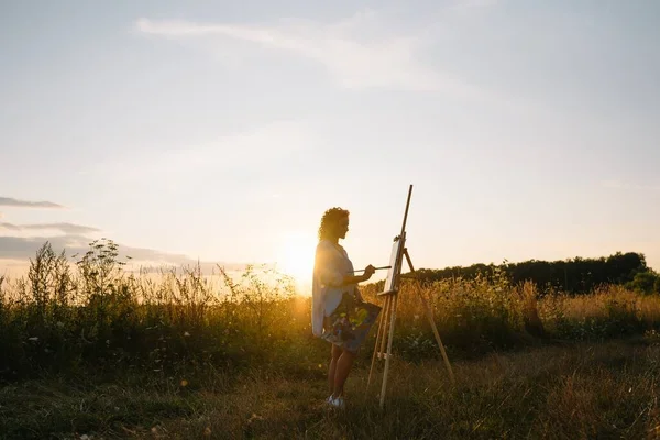 Silhouette of a blonde girl artist. Lady paints a painting on the canvas with the help of paints. A wooden easel keeps the picture. Summer is a sunny day, sunset