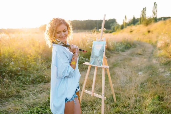 Pretty talented female painter painting on easel, making colorful sketches, creating marine landscape. Beautiful female artist painting with watercolor paints. Creativity and imagination concept.