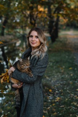Sonbahar parkında bir kız ve bir kedi. Kahverengi kazaklı bir kadın. Gri kedisiyle sonbahar parkında yürüyor.