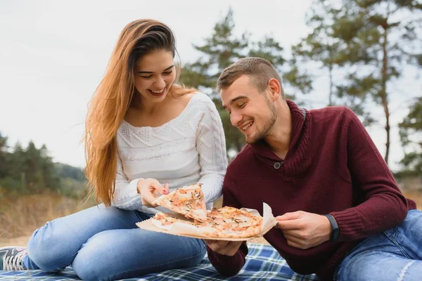 Young couple sitting on park bench and eating pizza.Fast food.Woman and man having picnic at sunset. Guys relaxing and enjoying food. Lifestyle