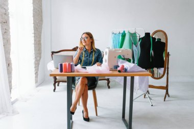 Gülümseyen moda tasarımcısı iş yerinde kameraya bakıyor, terzi, nakış işçisi ya da terzi masasında oturuyor renk kumaşları pantonu ve nakış desenleri ile duvarda, portre