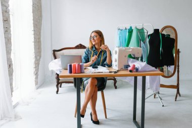 Gülümseyen moda tasarımcısı iş yerinde kameraya bakıyor, terzi, nakış işçisi ya da terzi masasında oturuyor renk kumaşları pantonu ve nakış desenleri ile duvarda, portre