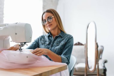 Gülümseyen moda tasarımcısı iş yerinde kameraya bakıyor, terzi, nakış işçisi ya da terzi masasında oturuyor renk kumaşları pantonu ve nakış desenleri ile duvarda, portre