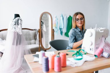 Gülümseyen moda tasarımcısı iş yerinde kameraya bakıyor, terzi, nakış işçisi ya da terzi masasında oturuyor renk kumaşları pantonu ve nakış desenleri ile duvarda, portre