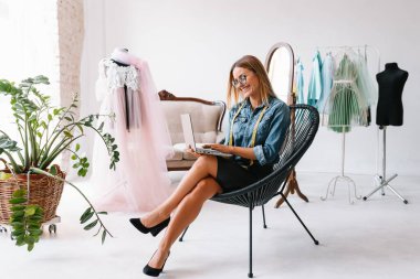 Gülümseyen moda tasarımcısı iş yerinde kameraya bakıyor, terzi, nakış işçisi ya da terzi masasında oturuyor renk kumaşları pantonu ve nakış desenleri ile duvarda, portre