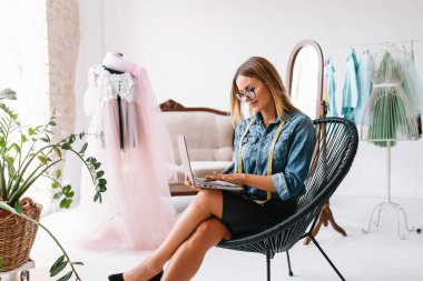 Gülümseyen moda tasarımcısı iş yerinde kameraya bakıyor, terzi, nakış işçisi ya da terzi masasında oturuyor renk kumaşları pantonu ve nakış desenleri ile duvarda, portre