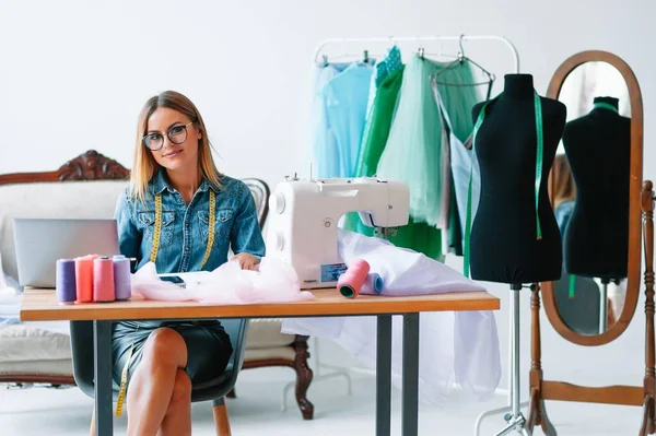 Gülümseyen moda tasarımcısı iş yerinde kameraya bakıyor, terzi, nakış işçisi ya da terzi masasında oturuyor renk kumaşları pantonu ve nakış desenleri ile duvarda, portre