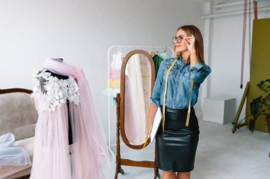 Gülümseyen moda tasarımcısı iş yerinde kameraya bakıyor, terzi, nakış işçisi ya da terzi masasında oturuyor renk kumaşları pantonu ve nakış desenleri ile duvarda, portre
