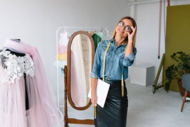 Gülümseyen moda tasarımcısı iş yerinde kameraya bakıyor, terzi, nakış işçisi ya da terzi masasında oturuyor renk kumaşları pantonu ve nakış desenleri ile duvarda, portre