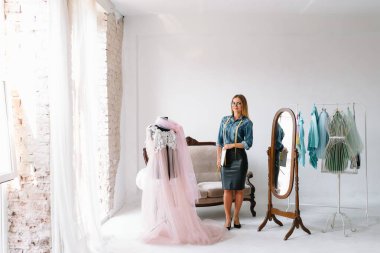 Gülümseyen moda tasarımcısı iş yerinde kameraya bakıyor, terzi, nakış işçisi ya da terzi masasında oturuyor renk kumaşları pantonu ve nakış desenleri ile duvarda, portre