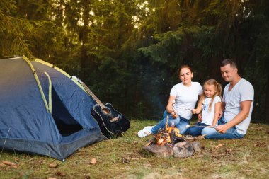 Üç kişilik bir aile kamp yapıyor ve ateşte yemek pişiriyor.