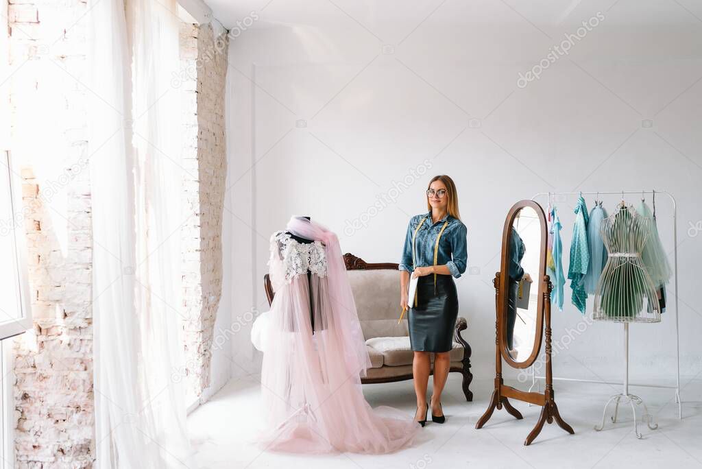 Smiling fashion designer looking at camera at workplace, dressmaker, needlewoman or tailor shop owner sitting at desk with color swatches pantone and embroidery design sketches on the wall, portrait