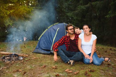 Üç kişilik bir aile kamp yapıyor ve ateşte yemek pişiriyor.