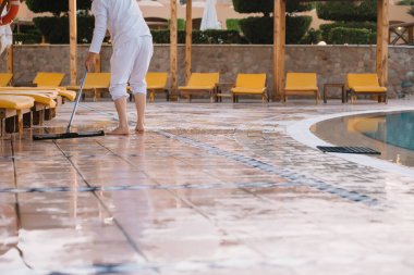Çalışan bir adam otelin bölgesindeki çöpleri topluyor ve havuzun yanındaki zemini yıkıyor.