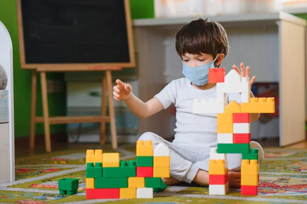 Little sad thoughtful bored toddler boy in mask playing colorful building blocks alone at home during quarantine. development game. Loneliness at self isolation period