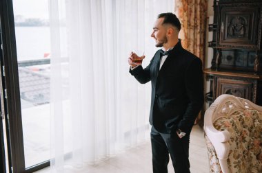 Şarap kadehli takım elbiseli şık bir adam, klasik odada duruyor. Moda..