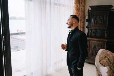 Şarap kadehli takım elbiseli şık bir adam, klasik odada duruyor. Moda..