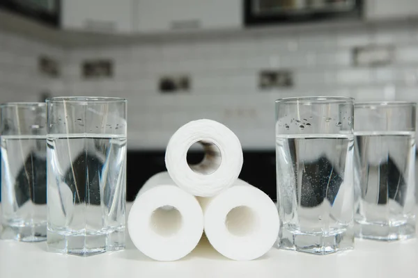 A glass of clean water with osmosis filter and cartridges on white table in a kitchen interior. Concept Household filtration system.