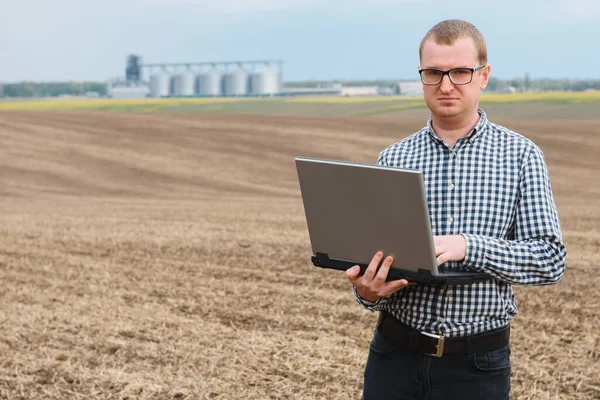 modern farmer checking his field plant and working on laptop computer against corn dryer silos in concept of industrial and agriculture